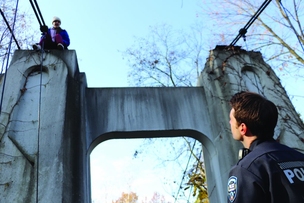 Officer stands below a concrete bridge arch while another person sits above secured by ropes, with cables, harness lines, bare trees, and blue sky in daylight