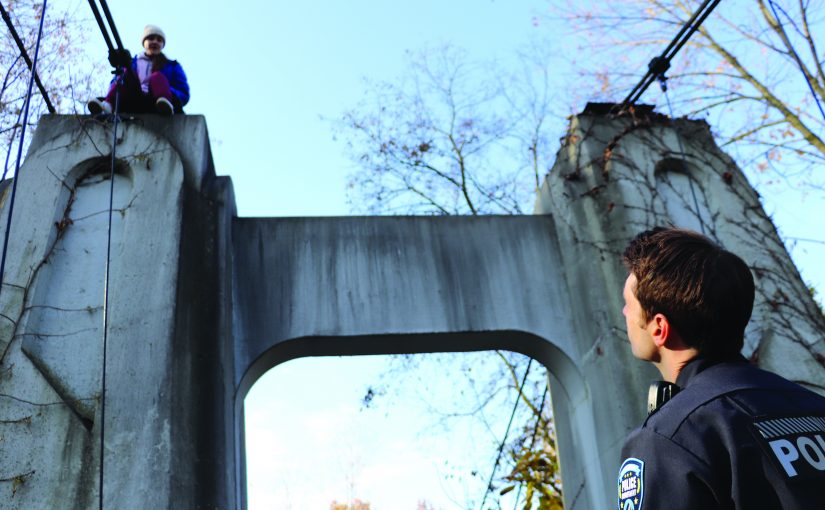 Officer stands below a concrete bridge arch while another person sits above secured by ropes, with cables, harness lines, bare trees, and blue sky in daylight