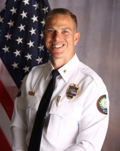 Police officer in white dress uniform with gold badge, black tie, and shoulder insignia, standing before a U.S. flag on a gray studio backdrop