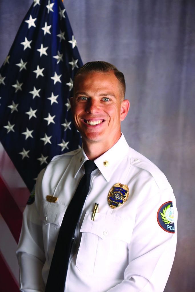 Uniformed police officer in white dress shirt with black tie stands before U.S. flag; visible badge, collar insignia, and shoulder patch