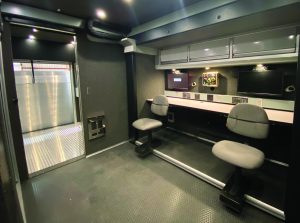 Inside mobile command center with swivel chairs, long desk, multiple monitors, overhead cabinets, and doorway to rear compartment.