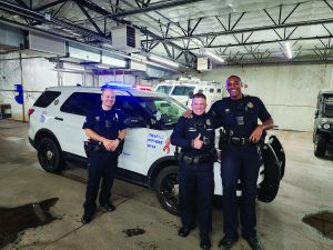 Three uniformed officers stand before marked police SUV with flashing lights in garage; other emergency vehicles visible in background, showing readiness