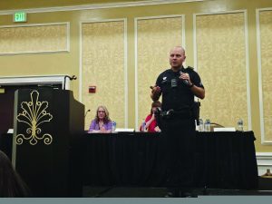 Police officer in uniform speaking at a podium during a traffic safety summit, holding a microphone with presentation materials and panelists seated behind
