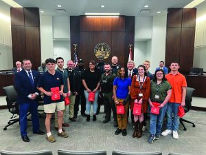 Group in courtroom holding red gift bags; behind them are city seal, American and state flags, wood paneling, and council seating area.