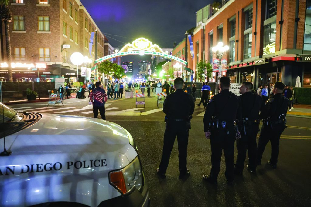 The image depicts a nighttime scene in an urban area, likely a downtown district. In the foreground, five police officers stand with their backs to the camera, observing the scene ahead. They are dressed in dark uniforms and utility belts. To the left of the officers is a San Diego Police vehicle with its headlights on and "San Diego Police" written on its side. The background features a well-lit street with several buildings lining both sides, including establishments like "Nobu" and other businesses. A large illuminated archway spans across the street in the distance, adorned with lights and signage that appears to welcome visitors to this part of town. Several pedestrians are visible under and around the archway, some interacting or walking by barricades set up in the middle of the street. The overall atmosphere suggests an event or situation requiring police presence for crowd control or public safety
