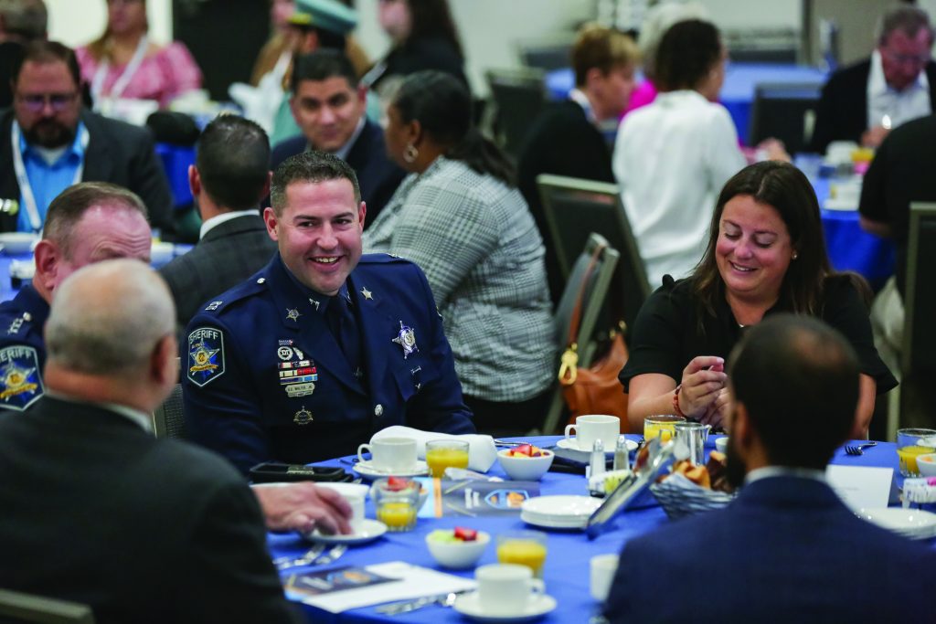 The image captures a formal event attended by law enforcement professionals. The setting is an indoor conference or banquet hall with multiple round tables draped in blue tablecloths. Each table is set with plates containing various food items, including fruit cups and pastries, along with cups of coffee or tea. Attendees are seated around the tables, engaged in conversation and enjoying their meals. Some individuals are dressed in uniforms featuring badges and insignia, while others are in civilian attire. The background shows more attendees and a blurred view of the hall, indicating a well-attended gathering