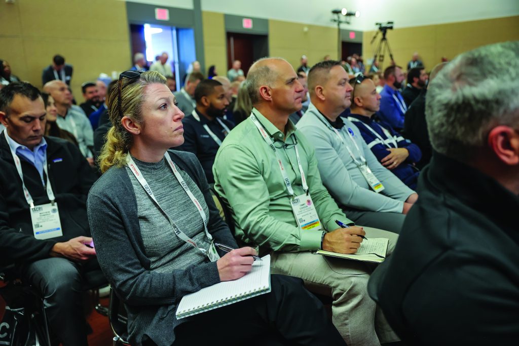 Large group of law enforcement professionals seated in rows at conference, taking notes with pens and notebooks; visible lanyards with badges