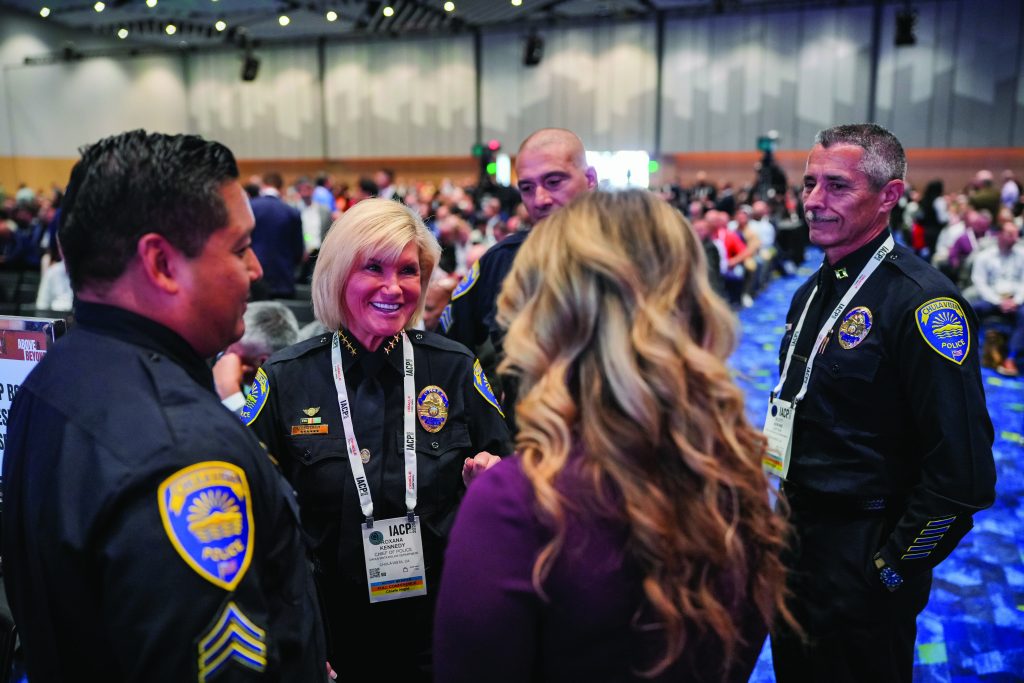 Group of uniformed police officers with conference badges talking to an attendee in a large event hall with blue patterned carpet and rows of seated audience