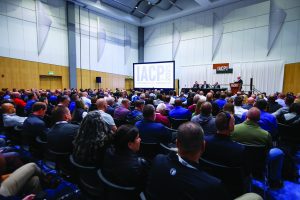 Attendees fill a large conference hall facing a stage with panelists, an IACP 2025 presentation screen, signage, and room lighting panels overhead
