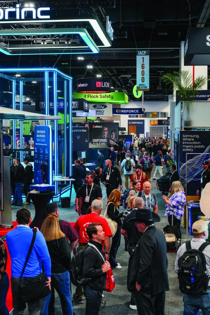 Busy law enforcement trade show floor with attendees walking among booths featuring technology displays, signage for safety solutions, and vendor exhibits