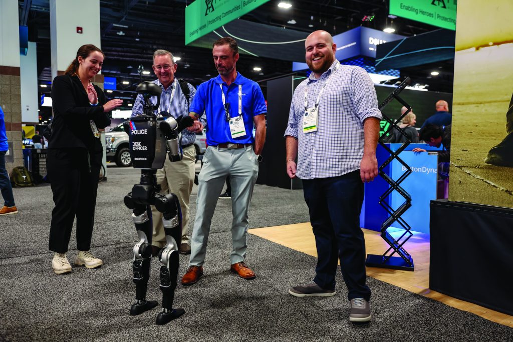 Four people standing on a trade show floor observing a humanoid police robot labeled ‘Police The Officer,’ with tech booths and banners visible in the background