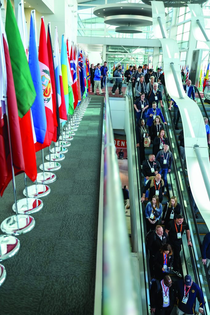 Large convention center with rows of international flags on the left and attendees riding escalators on the right, bright natural light streaming through glass walls