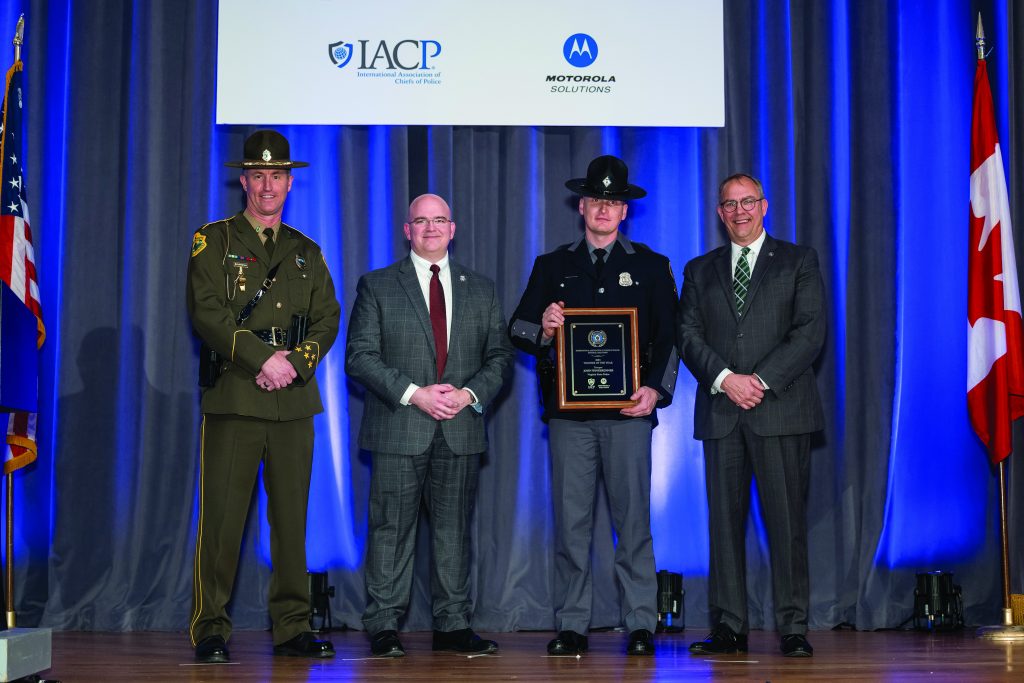 Four uniformed and suited individuals stand on a stage holding an award plaque beneath IACP and Motorola logos, with U.S. and Canadian flags and blue curtains behind