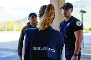 Three uniformed law enforcement professionals, one in Europol vest, stand outdoors with trees and mountain landscape in background
