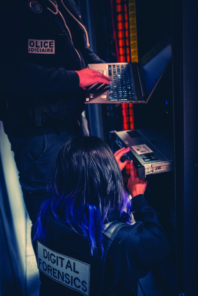 Two officers labeled "POLICE JUDICIAIRE" and "DIGITAL FORENSICS" analyze data on a laptop and server in a dim room lit by orange ambient lighting
