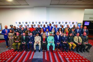 Large group of law enforcement professionals posed indoors in two rows, wearing formal uniforms and suits, holding awards in front of a backdrop with IACP 40 Under 40 logos.