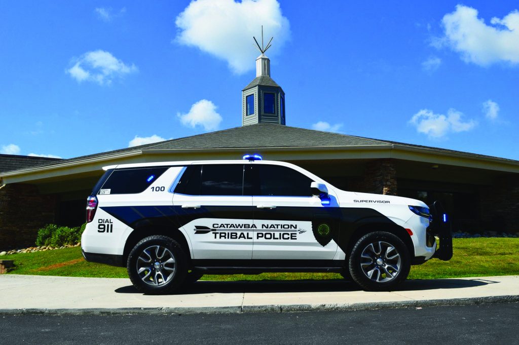 Marked Catawba Nation Tribal Police SUV with blue lights is parked on grass beside a brick building with cupola, flagpole, and clear sky above