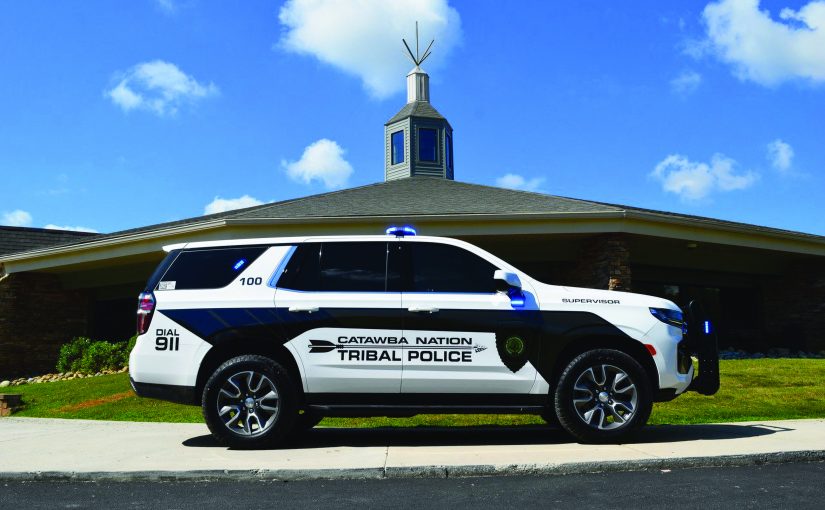 Marked Catawba Nation Tribal Police SUV with blue lights is parked on grass beside a brick building with cupola, flagpole, and clear sky above