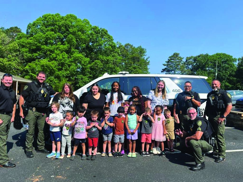 Uniformed officers and adults stand with a line of children in front of a marked patrol SUV, outdoors on pavement with trees, buildings, and clear blue sky behind