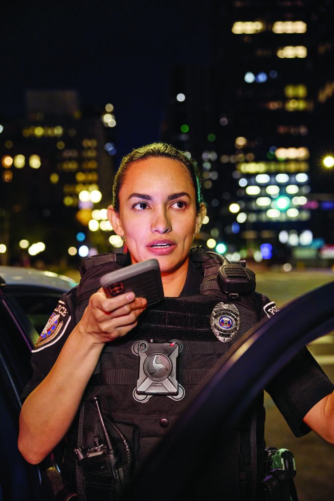 Uniformed officer in body armor holds a handheld radio beside a patrol vehicle at night, with badge, camera, shoulder patches, and blurred city lights in the background