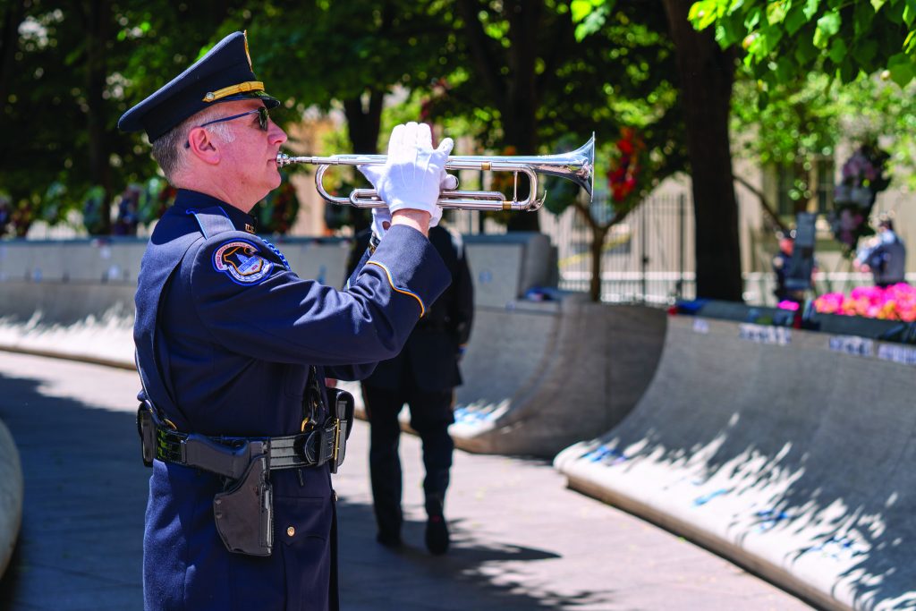 A uniformed law enforcement officer stands outdoors in a park-like setting, playing a silver trumpet. The officer is wearing a dark blue dress uniform with white gloves, a peaked cap, and has a holstered firearm on the right hip. The officer's face is blurred for anonymity. In the background, there are trees with green leaves providing shade and sunlight filtering through. There is also a curved stone wall adorned with small blue and white flags or decorations along its top edge. Another person in uniform can be seen in the distance, partially obscured by shadows and trees. The scene appears to be part of a formal ceremony or memorial event.