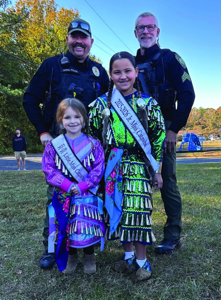 Two uniformed officers stand behind two children wearing sashes reading Little Miss Catawba and Jr Miss Catawba, outdoors near road, trees and playground