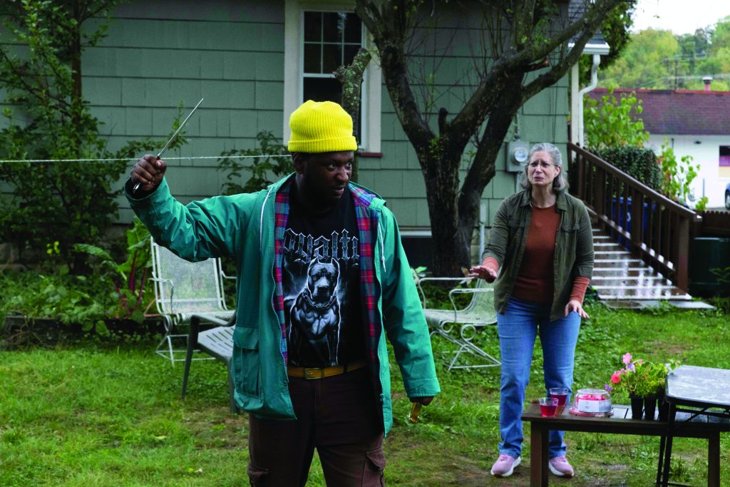 Individual in yellow beanie and green jacket holds a knife in a backyard near a house as another person reaches forward, with patio chairs, table items, grass, and trees