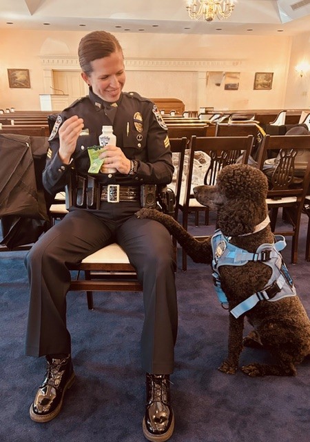 Woman in police uniform sitting on chair with bottled beverage and a black poodle in a therapy dog vest sitting next to her with its paw on her lap.