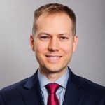 Person wearing a dark navy suit jacket, light blue dress shirt, and bright red tie against a plain gray background in a formal portrait pose