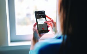 A person near a window holds a smartphone displaying an active 911 emergency call screen with a prompt to share live video, tapping the option on the display