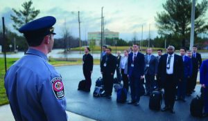 Uniformed officer faces group in formal attire standing in formation with luggage on ground outside building; scene suggests briefing or deployment