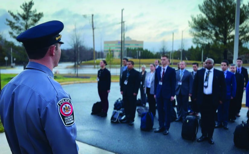 Uniformed officer faces group in formal attire standing in formation with luggage on ground outside building; scene suggests briefing or deployment