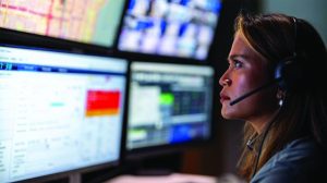 Communications operator wearing a headset monitors four screens displaying maps, call data, camera feeds, and software in a dim control room setting