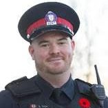Police officer in uniform with blurred face, black vest labeled 'POLICE,' red-black hat with emblem, and red poppy pin displayed on vest