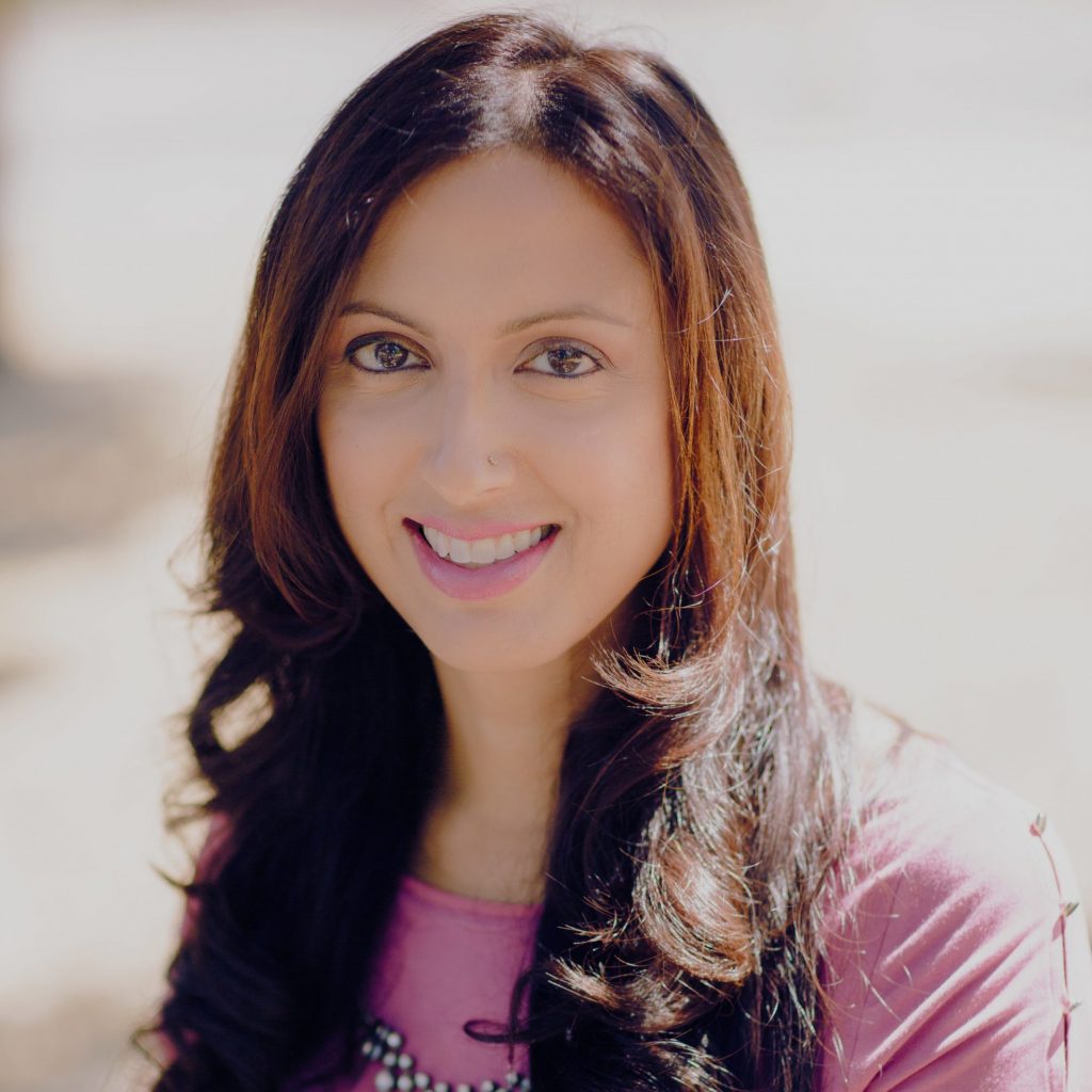 Portrait-style image of an individual wearing a pink short‑sleeve top and necklace, shown outdoors with long hair draped over shoulders and a softly blurred sunlit background