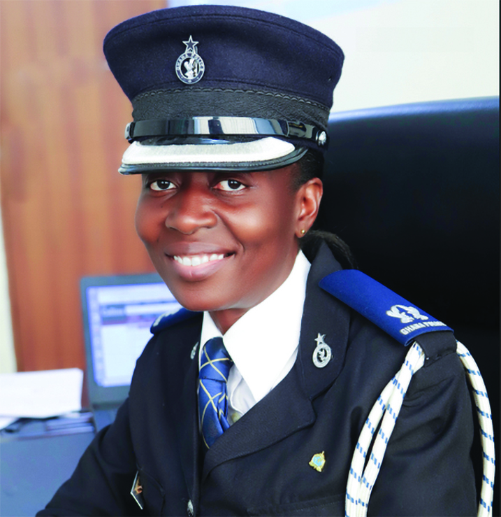 Uniformed law enforcement officer seated at a desk wearing a peaked cap, tie, shoulder cord, and badges, with a computer monitor and office equipment behind