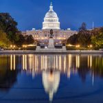 Night view of illuminated U.S. Capitol with dome reflection in water, framed by trees and lights under a clear blue sky, symbolizing governance.