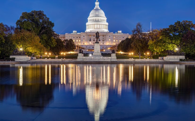 Night view of illuminated U.S. Capitol with dome reflection in water, framed by trees and lights under a clear blue sky, symbolizing governance.