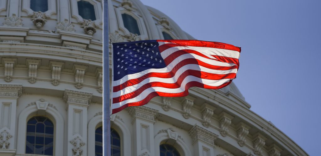 American flag waves before U.S. Capitol dome with ornate carvings, columns, and windows, set against a clear blue sky