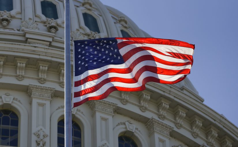 American flag waves before U.S. Capitol dome with ornate carvings, columns, and windows, set against a clear blue sky