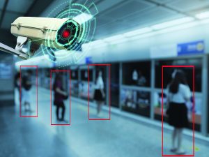 a surveillance camera mounted on the left side, focusing on a subway platform. The camera has a digital overlay with a red center and blue concentric circles, indicating it is actively recording or analyzing. There are four red rectangles highlighting individuals on the platform, suggesting they are being monitored or identified by the surveillance system. The background is slightly blurred, showing people waiting near the subway tracks and advertisements on the walls.