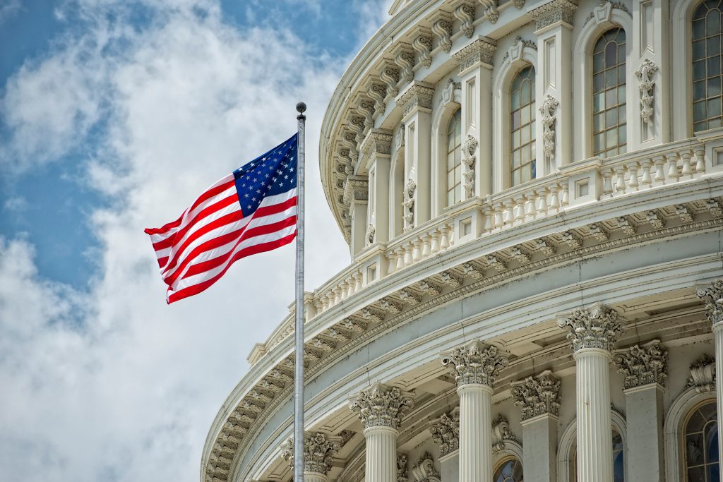 American flag waves before U.S. Capitol’s ornate facade with tall columns, arched windows, and partly cloudy sky
