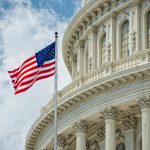American flag waves before U.S. Capitol’s ornate facade with tall columns, arched windows, and partly cloudy sky