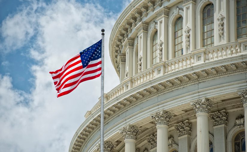 American flag waves before U.S. Capitol’s ornate facade with tall columns, arched windows, and partly cloudy sky