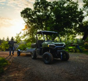 Green off-road utility vehicle with trailer parked on dirt path in wooded area at sunset; person nearby gestures toward the vehicle.