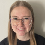 Individual with long, straight light brown hair wearing a black shirt, standing against a plain light-colored background