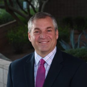 Individual in dark suit, pink tie, and checkered shirt stands outdoors before greenery and brick wall