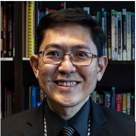 Individual wearing rectangular eyeglasses, dark suit jacket, patterned dress shirt, striped tie, and conference lanyard sits before bookshelves filled with books, journals, and reference materials, with dark shelving and colorful book spines visible