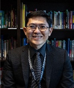 Person in dark suit and patterned tie sits before shelves filled with books in an office or library setting.