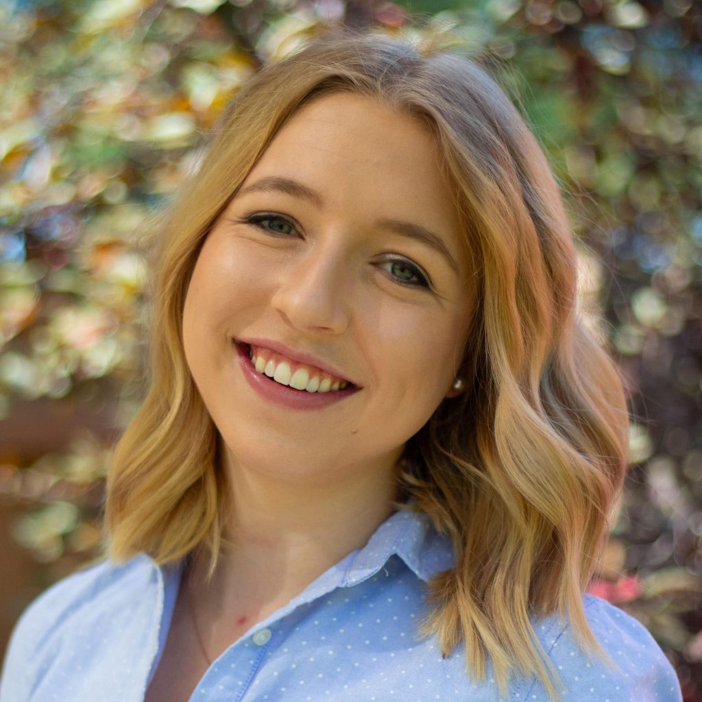 Individual with shoulder-length blonde hair in light blue polka-dot shirt stands outdoors; background shows blurred green foliage and sunlight